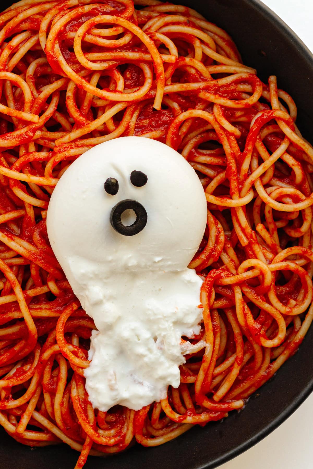 Spaghetti and tomato sauce on a black plate topped with a burrata ghost.