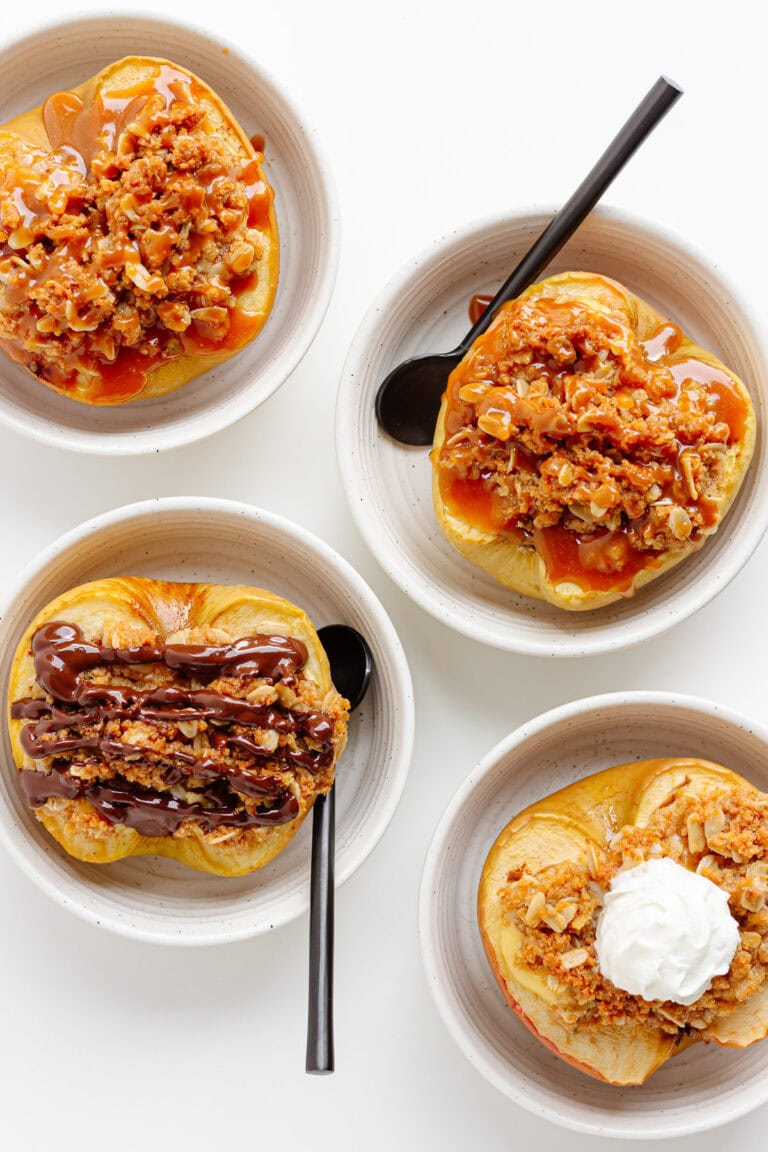 Four cheesecake stuffed baked apples in small serving bowls topped with various toppings.
