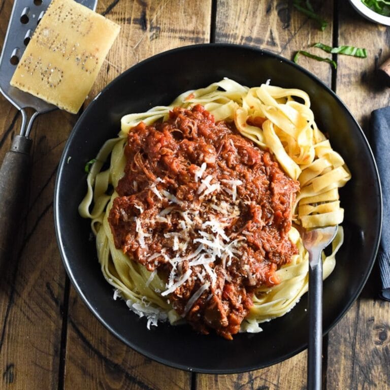 A black plate of turkey bolognese pasta with parmesan and a grater next to it.