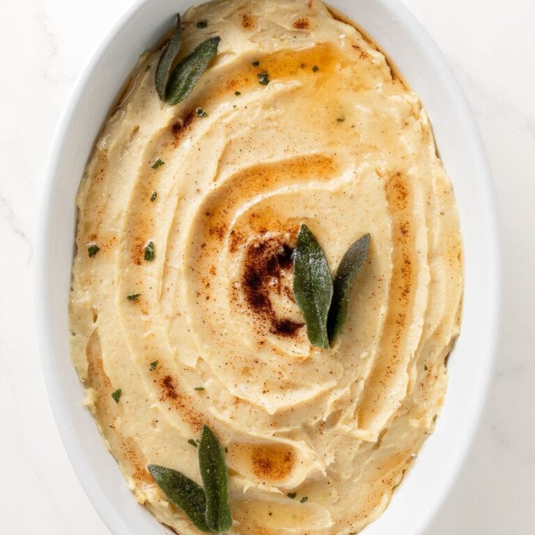 Mashed white sweet potatoes in a white oval serving dish drizzled with brown butter and garnished with sage leaves.