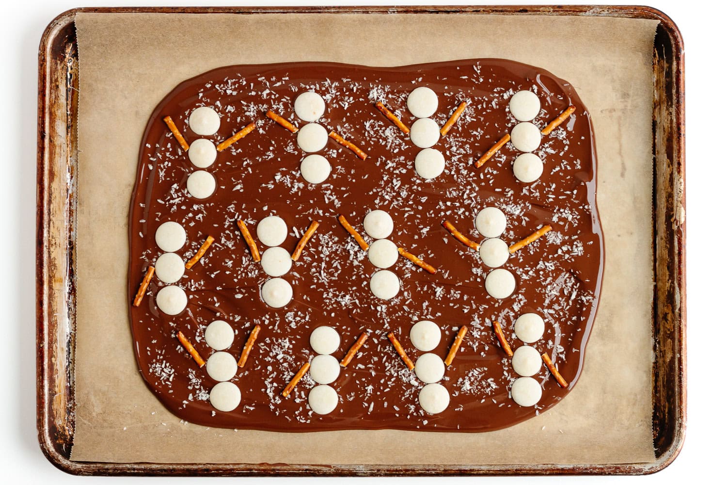 Snowmen shapes being assembled on chocolate bark with three white chocolate wafers and two pretzel stick arms per snowman.