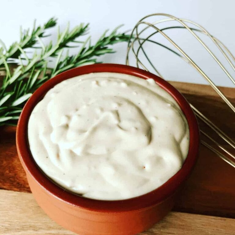 Creamy horseradish sauce in a brown bowl sitting on a wooden board next to a whisk and fresh herbs.