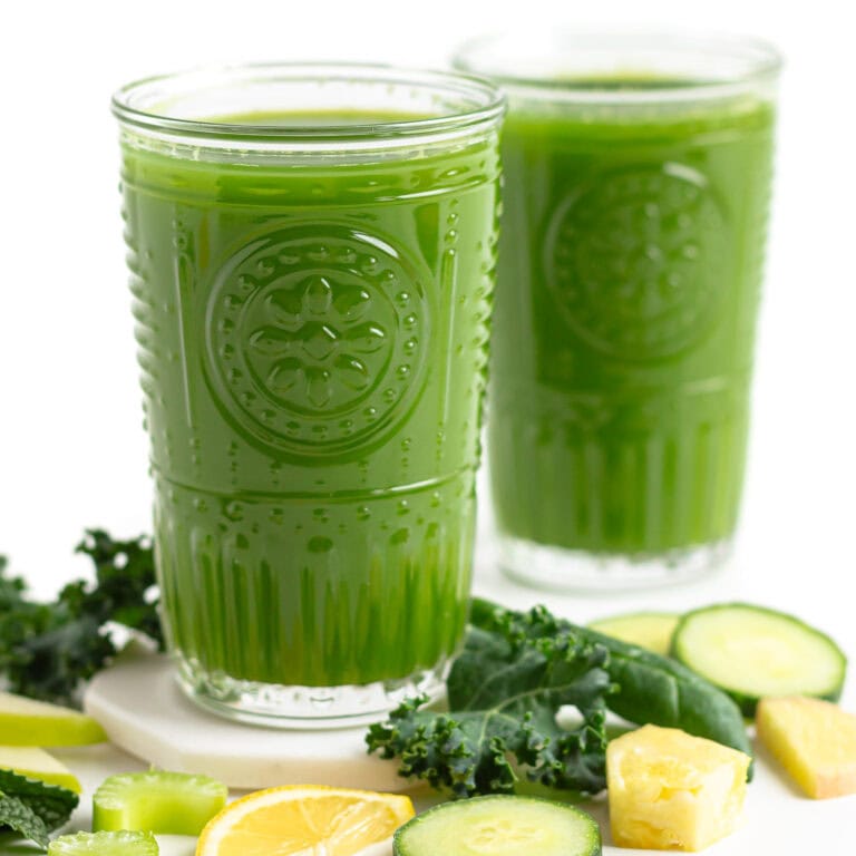 Two glasses of green blender juice surrounded by some of the ingredients.