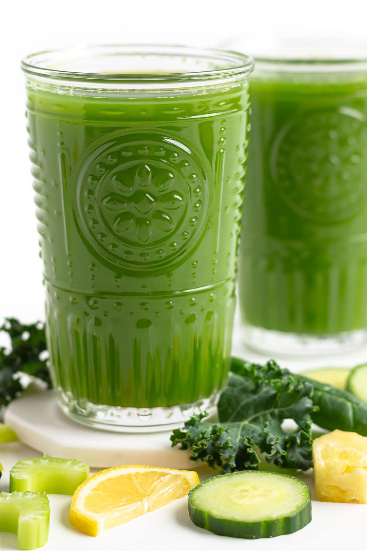 Two glasses of green juice with some vegetables and fruit scattered around them.
