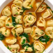 A bowl of beef tortellini soup with gold colored spoon.