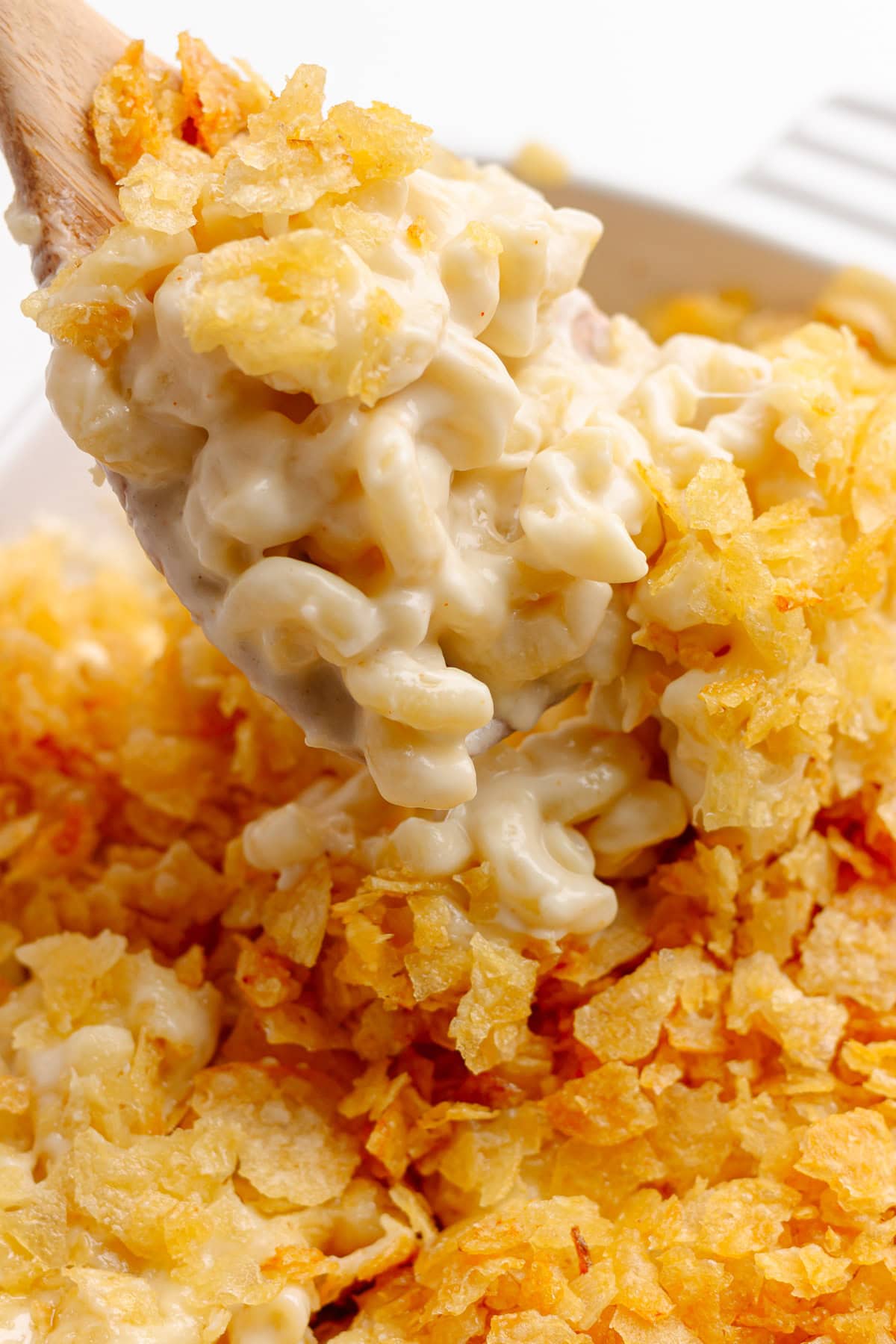 Wooden spoon lifting some baked white cheddar mac and cheese from a white casserole dish.