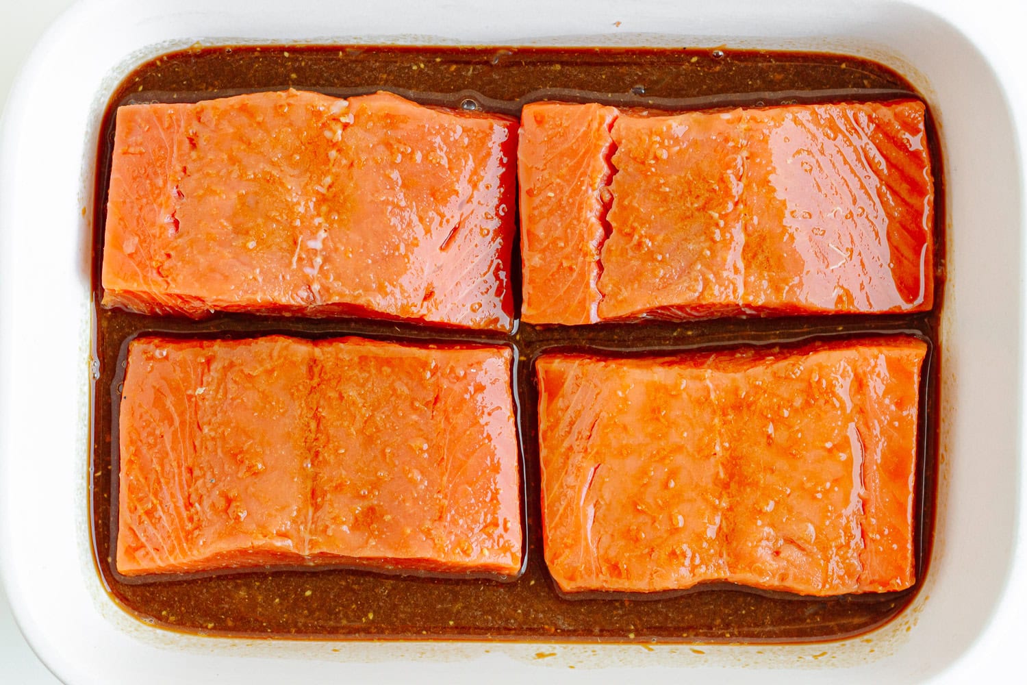 Four salmon filets marinating in a white ceramic dish.