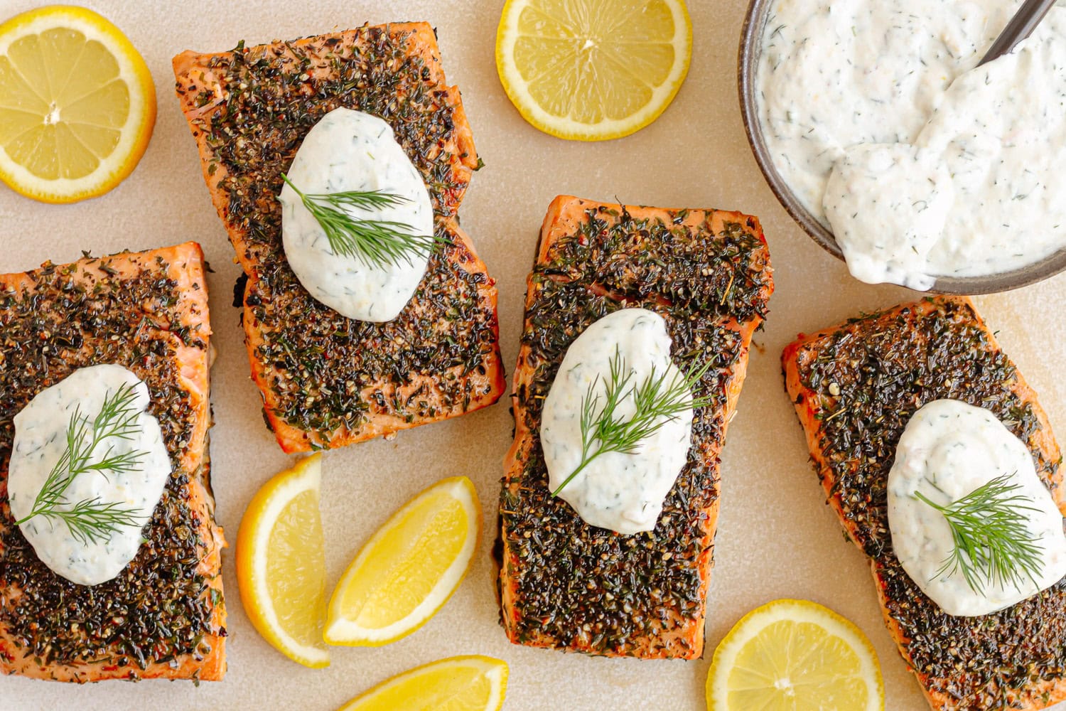 Herb crusted salmon filets with lemon dill sour cream sauce and lemon slices on a serving platter.