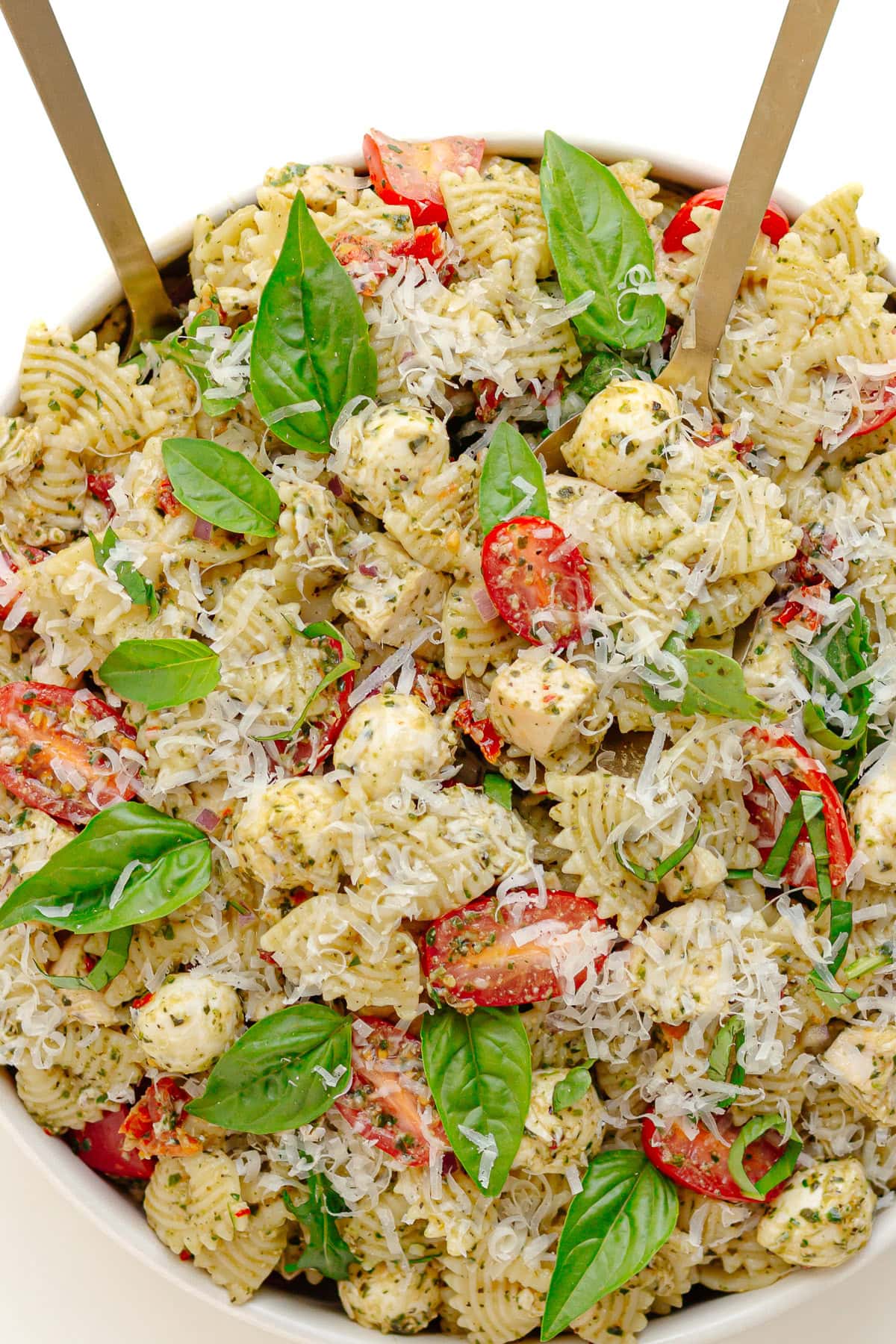 White serving bowl of pesto chicken pasta salad with two salad servers sticking out.
