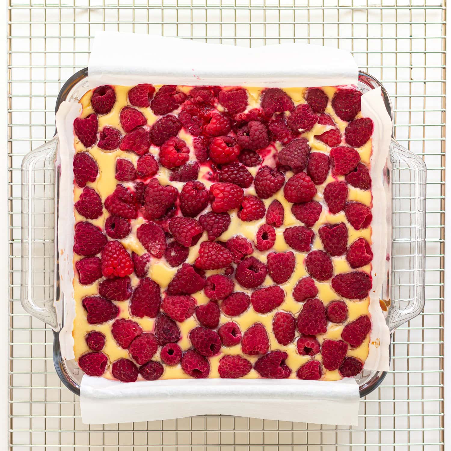 Baked raspberry cheesecake bars in glass baking dish on a wire cooling rack.