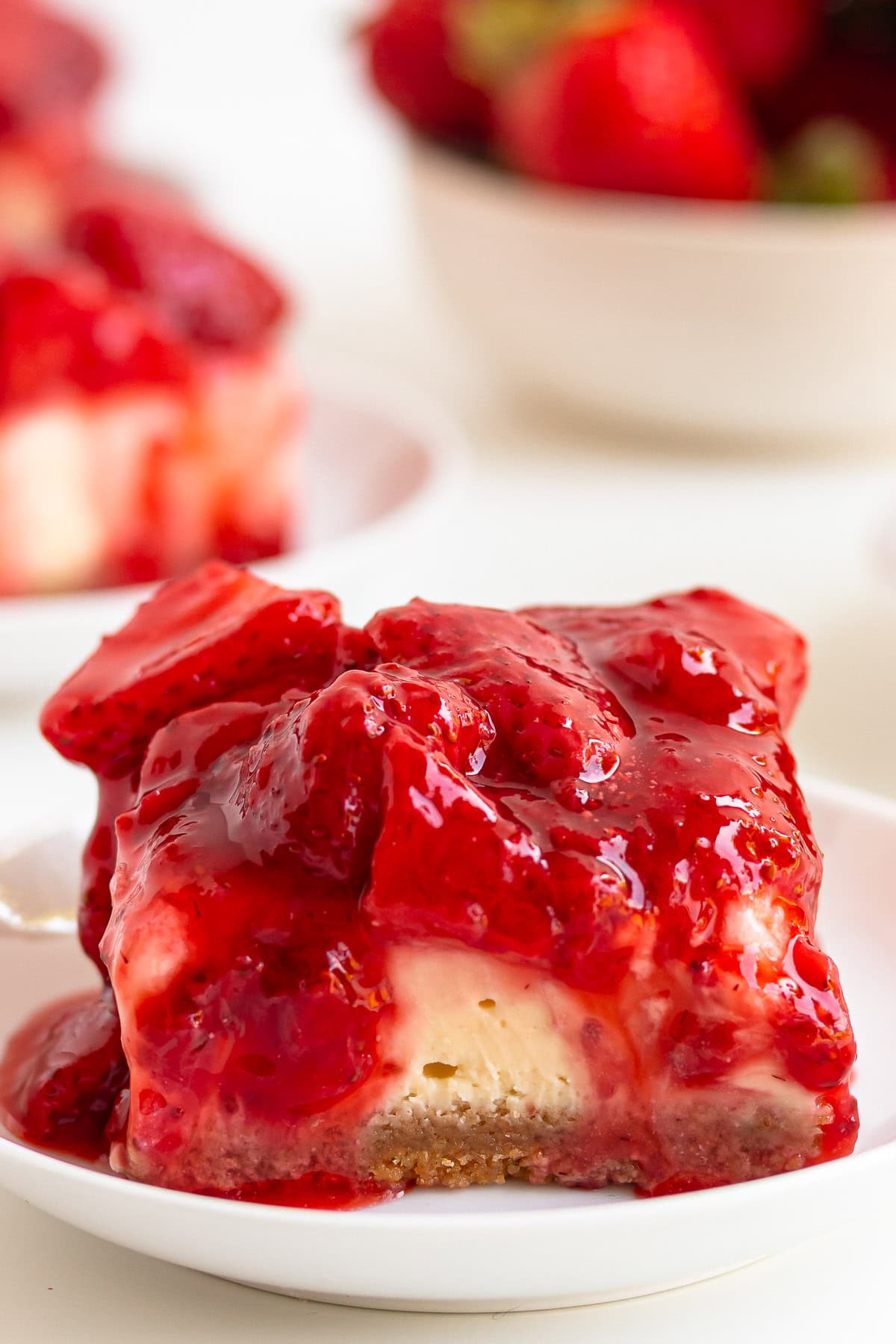 Strawberry cheesecake bar on a white plate with bowl of strawberries in the background.