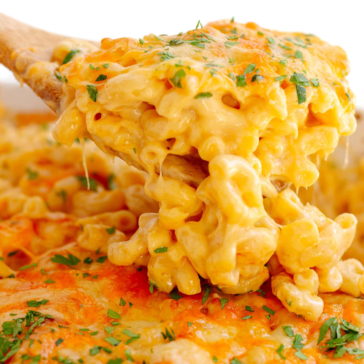 Wooden spoon lifting up cheesy baked mac and cheese from casserole dish.