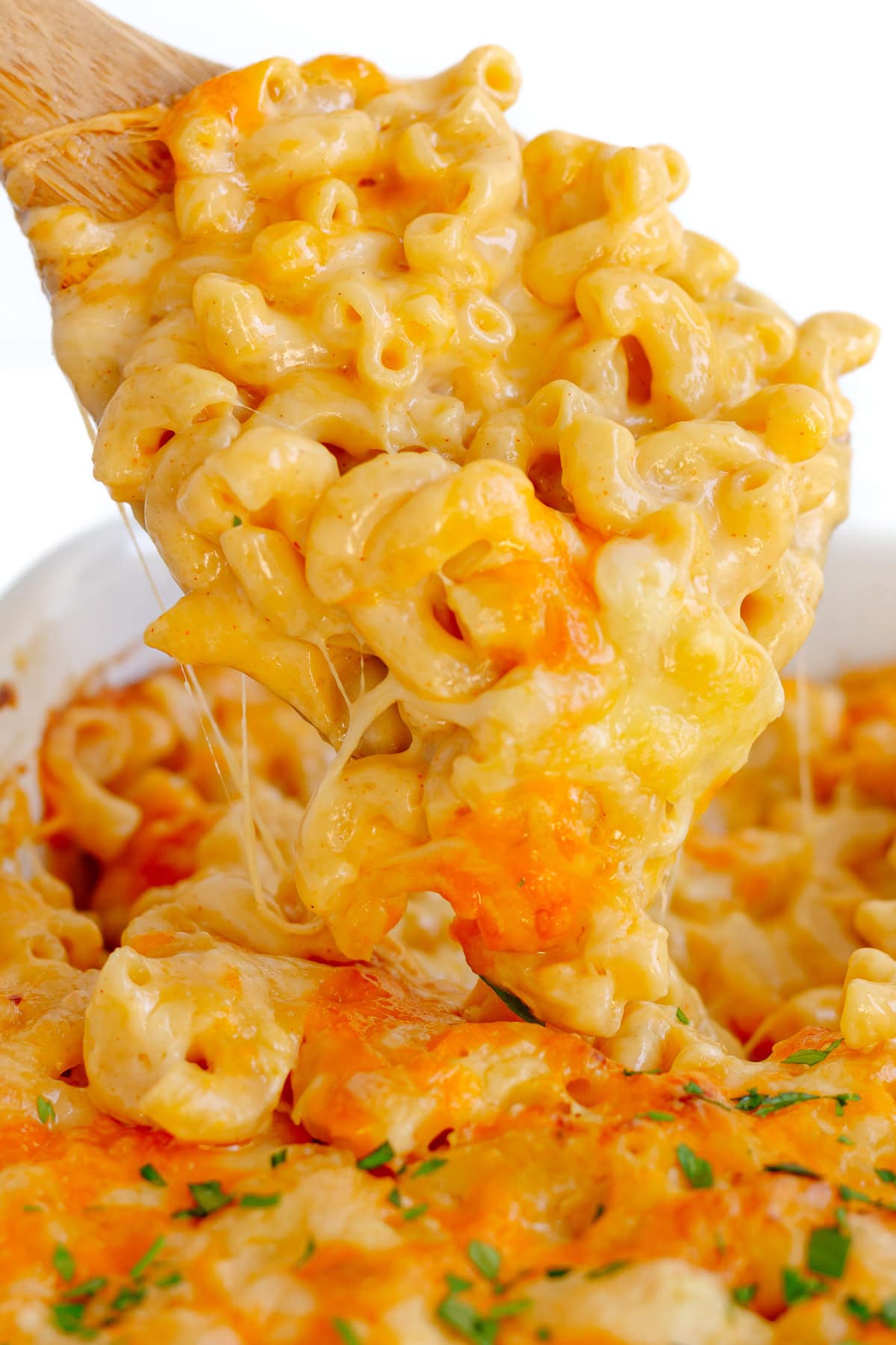 Wooden spoon lifting up some three cheese macaroni and cheese from baking dish.