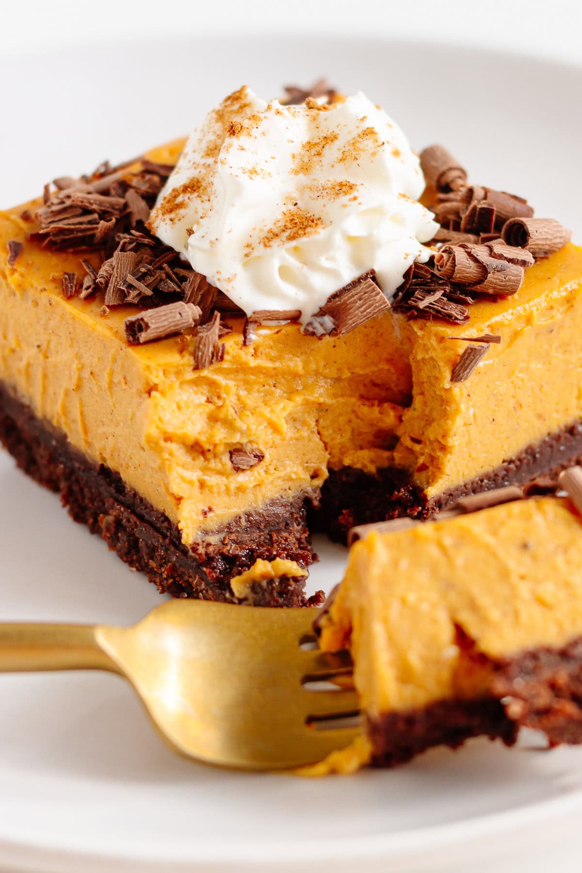 Pumpkin cheesecake bar on a white plate with gold colored fork scooping out a bite.