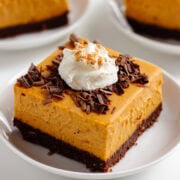 Pumpkin cheesecake bars on white serving plates garnished with chocolate shavings, whipped cream and pumpkin pie spice.