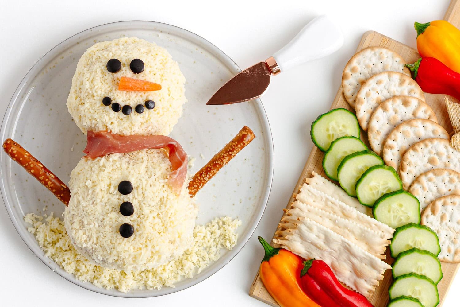 Snowman shaped cheese ball on a plate next to a board of crackers and raw veggies.
