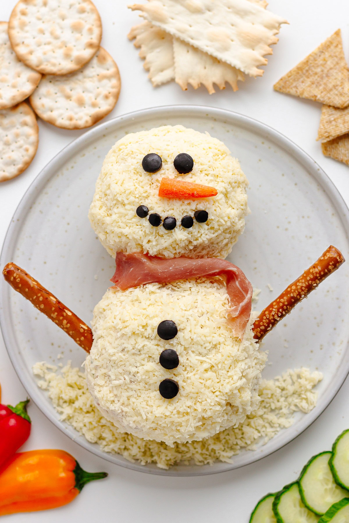 A snowman Christmas cheese ball on a plate surrounded by a variety of crackers and veggies.
