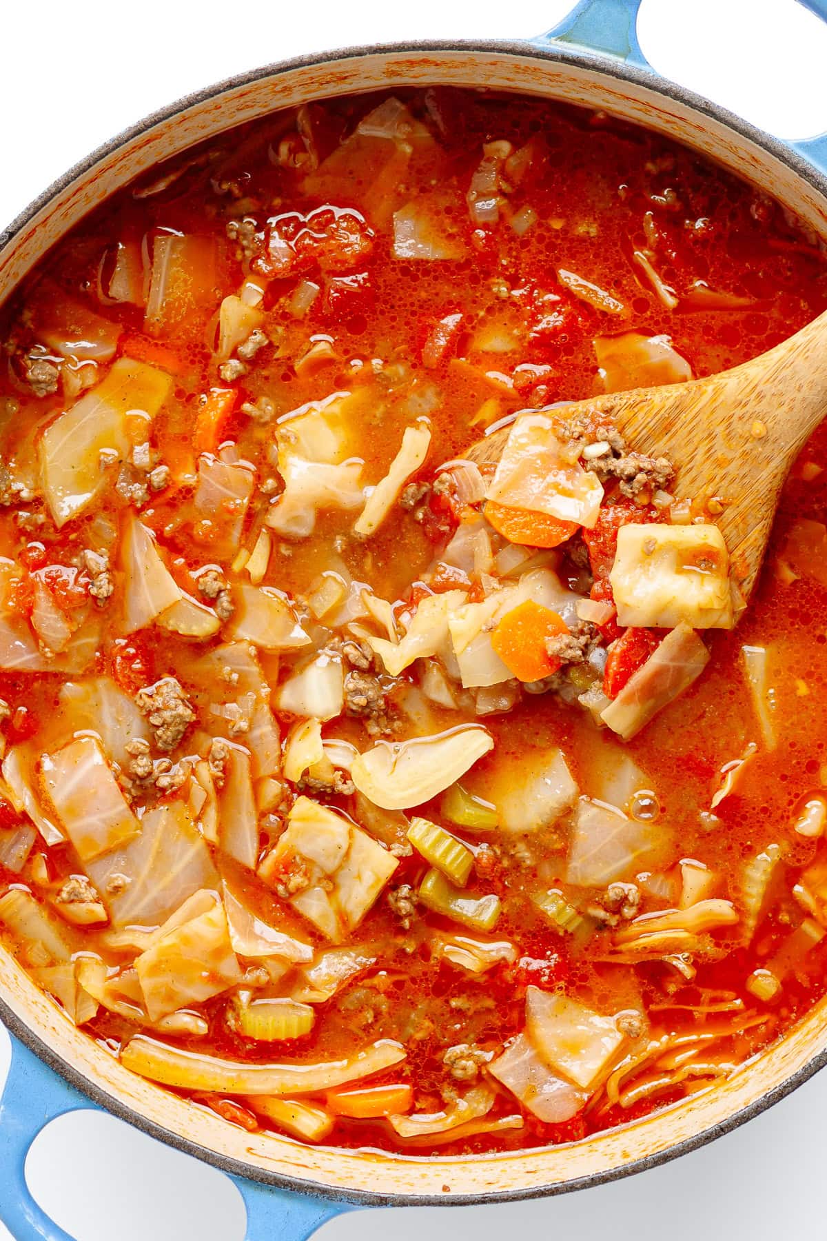 Wooden spoon stirring ground beef and cabbage soup in a Dutch oven.