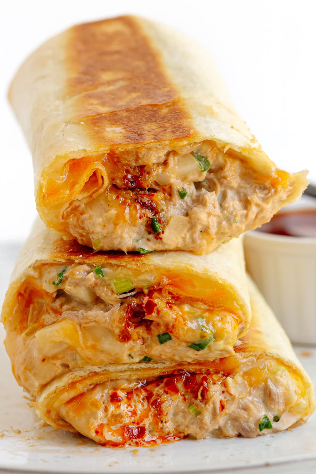 Spicy tuna melt wraps piled up on a plate next to a small bowl of chili crisp.