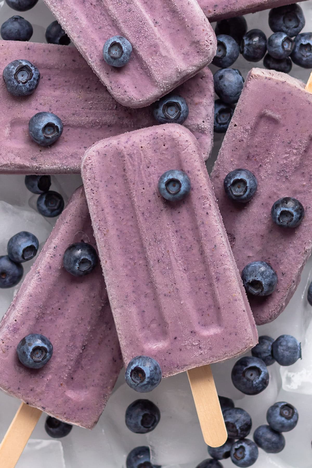 Creamy blueberry popsicles piled on a tray of ice with fresh blueberries.