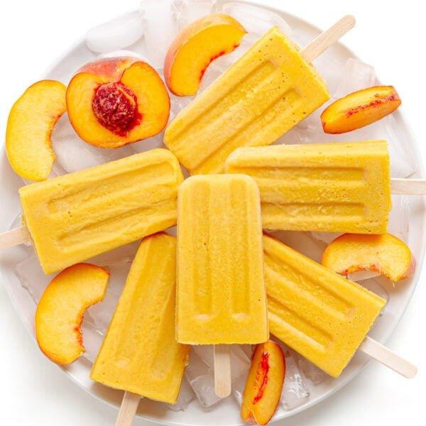 Creamy peach popsicles piled on a plate of ice with fresh peach slices.