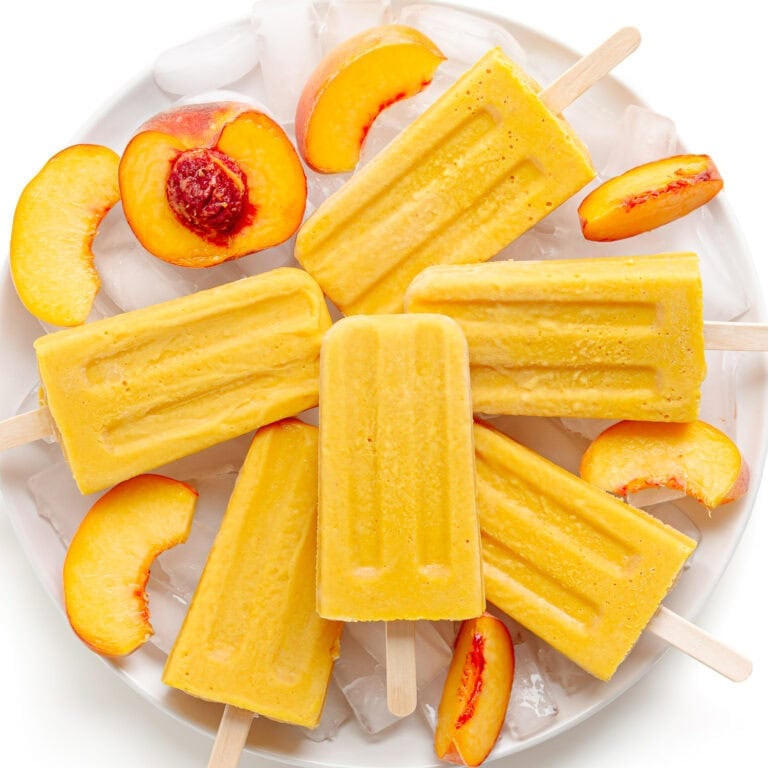 Creamy peach popsicles piled on a plate of ice with fresh peach slices.