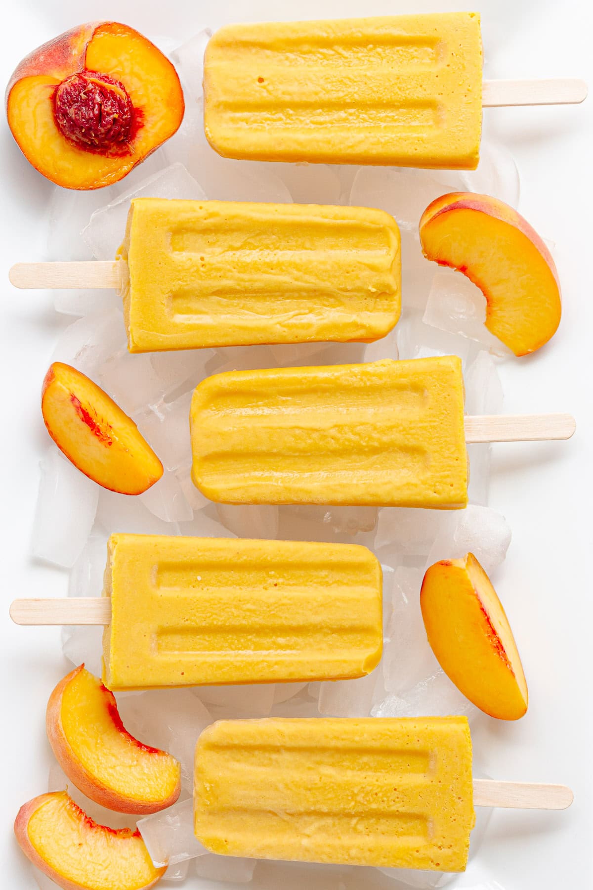 Peach popsicles lined up on a white tray of ice with fresh peach slices.