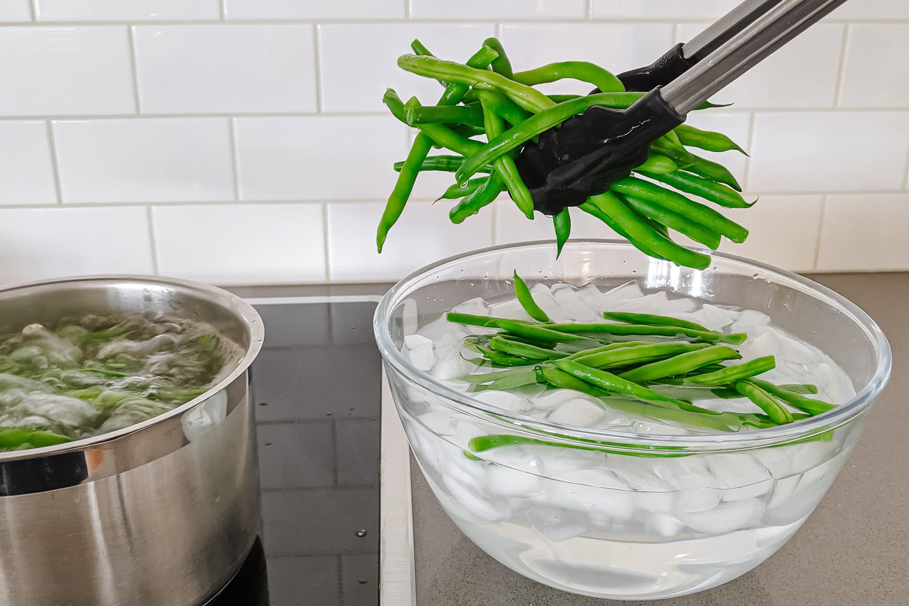 Tongs pulling green beans from pot of boiling water and placing into bowl of ice water.