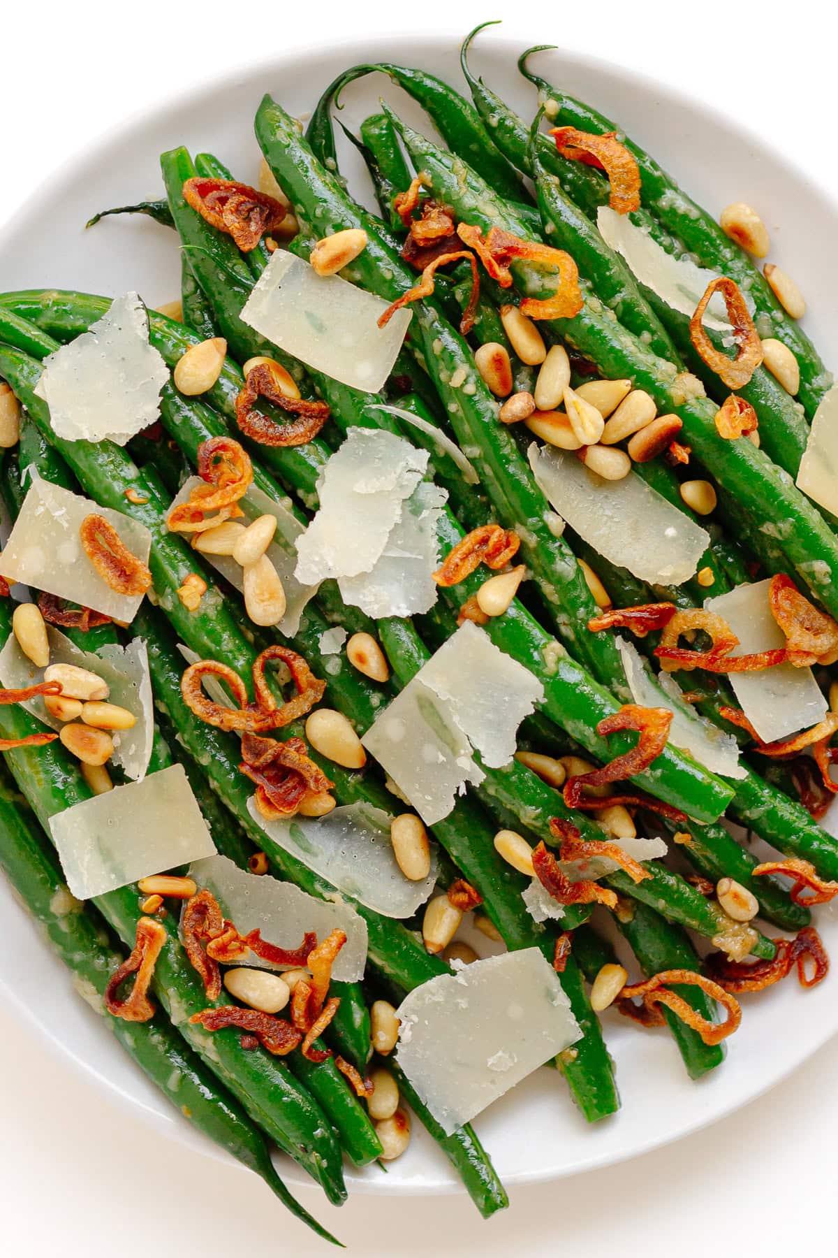 Garlic parmesan green beans on a white serving plate garnished with pine nuts, parmesan shavings and fried shallots.
