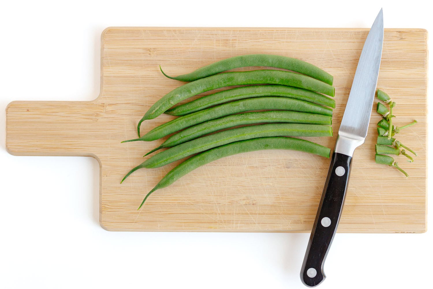 Green beans lined up on a wood cutting board with the stems trimmed off by a paring knife.